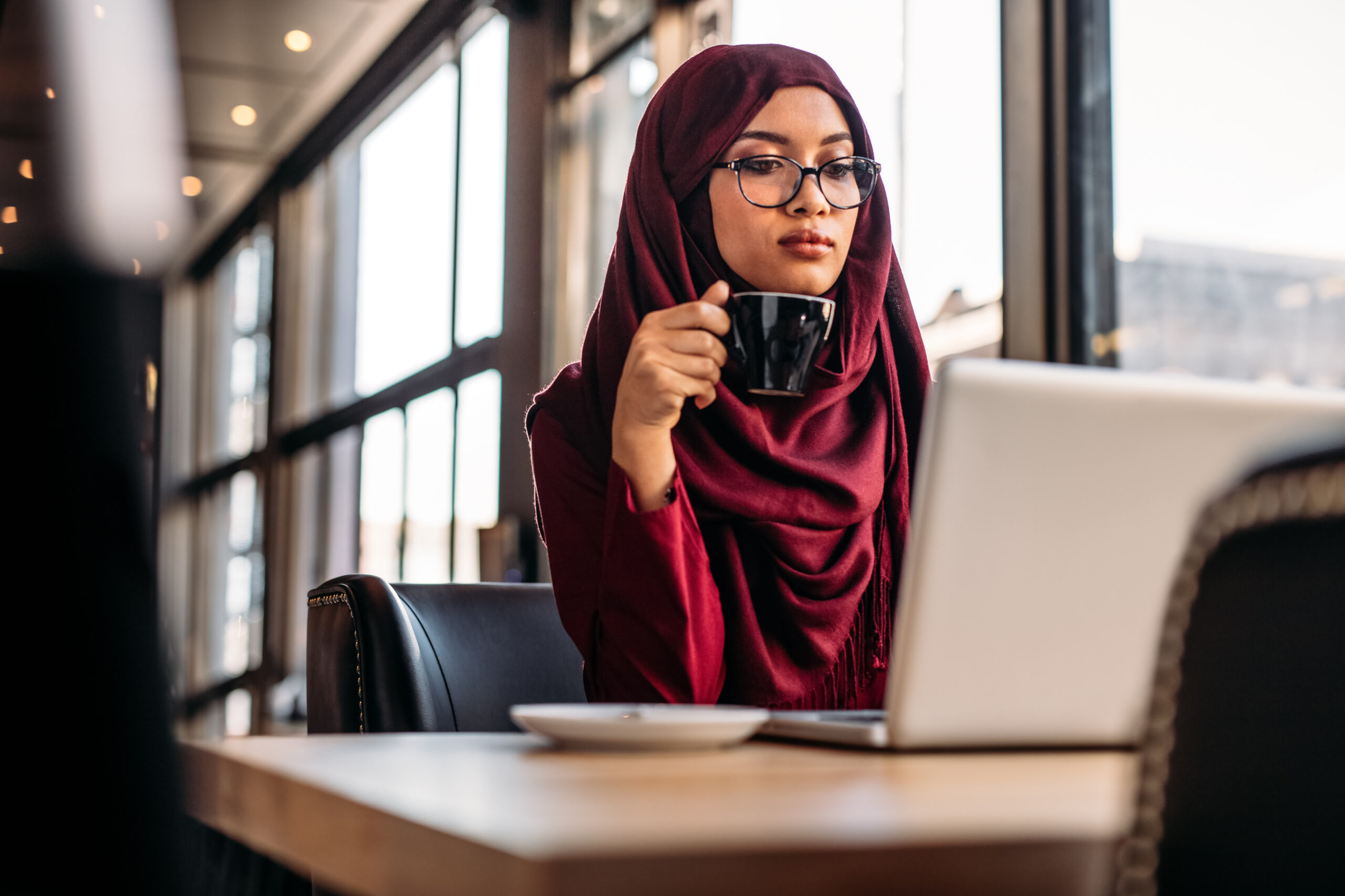 Muslim woman working at a cafe and using laptop computer. Female in hijab drinking coffee and looking at laptop in table at restaurant.