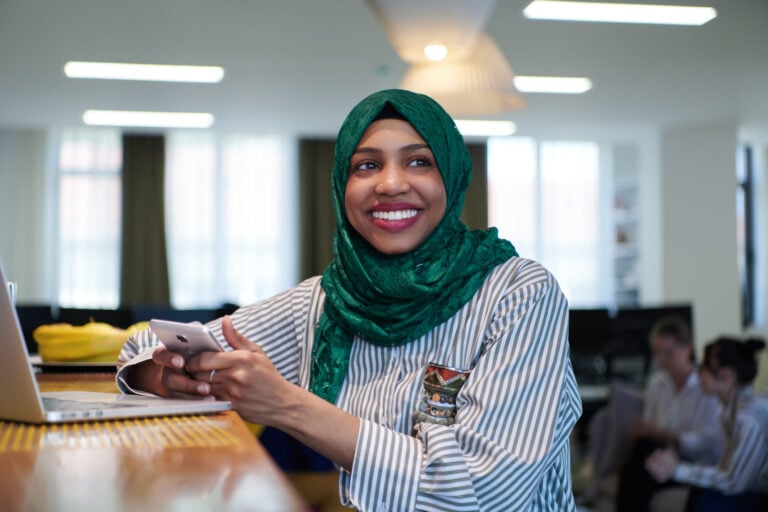 African muslim business woman wearing a green hijab using mobile phone while working on laptop computer in relaxation area at modern open plan startup office.