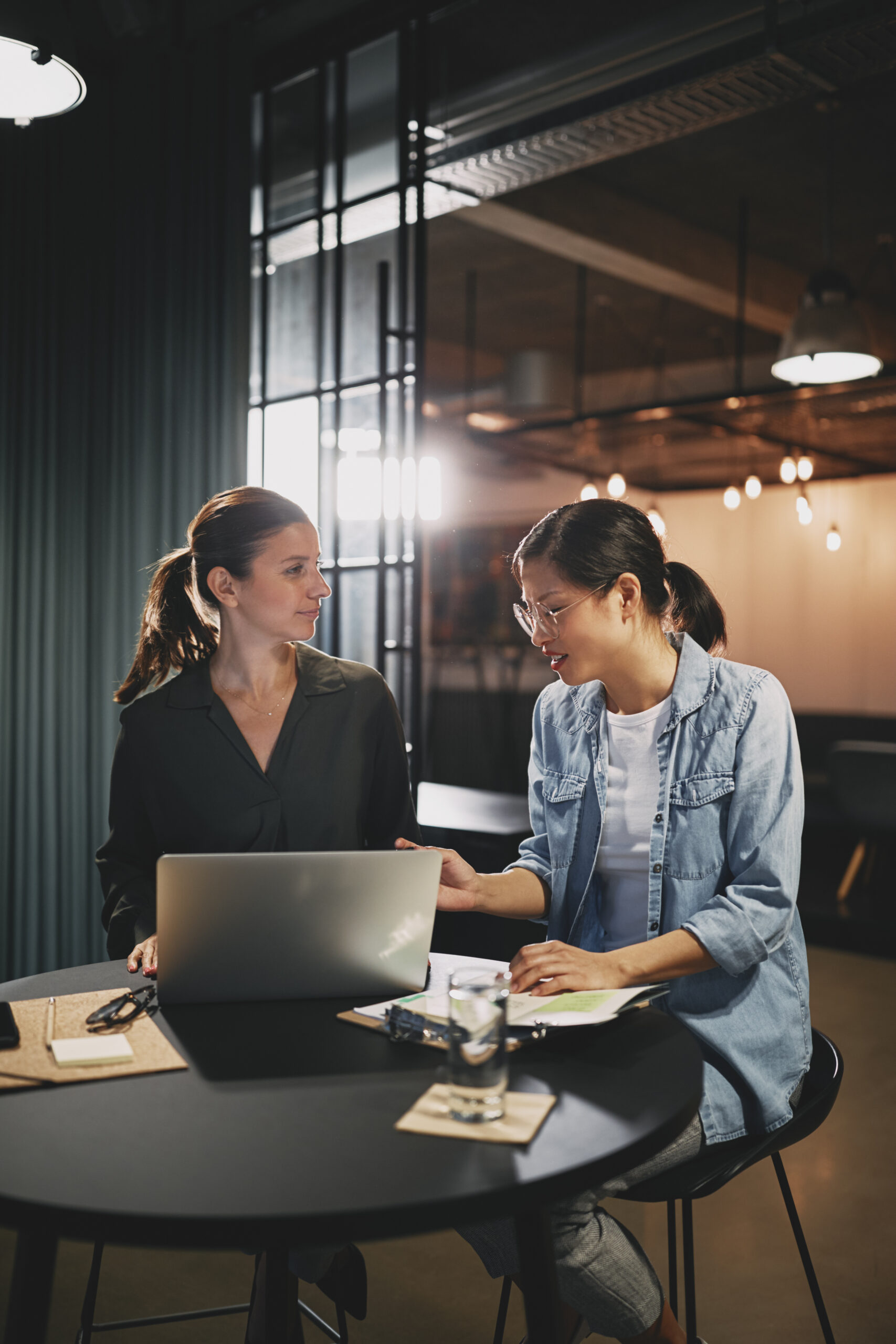 Two young businesswomen sitting at a table in a modern office working on a laptop and going over paperwork.