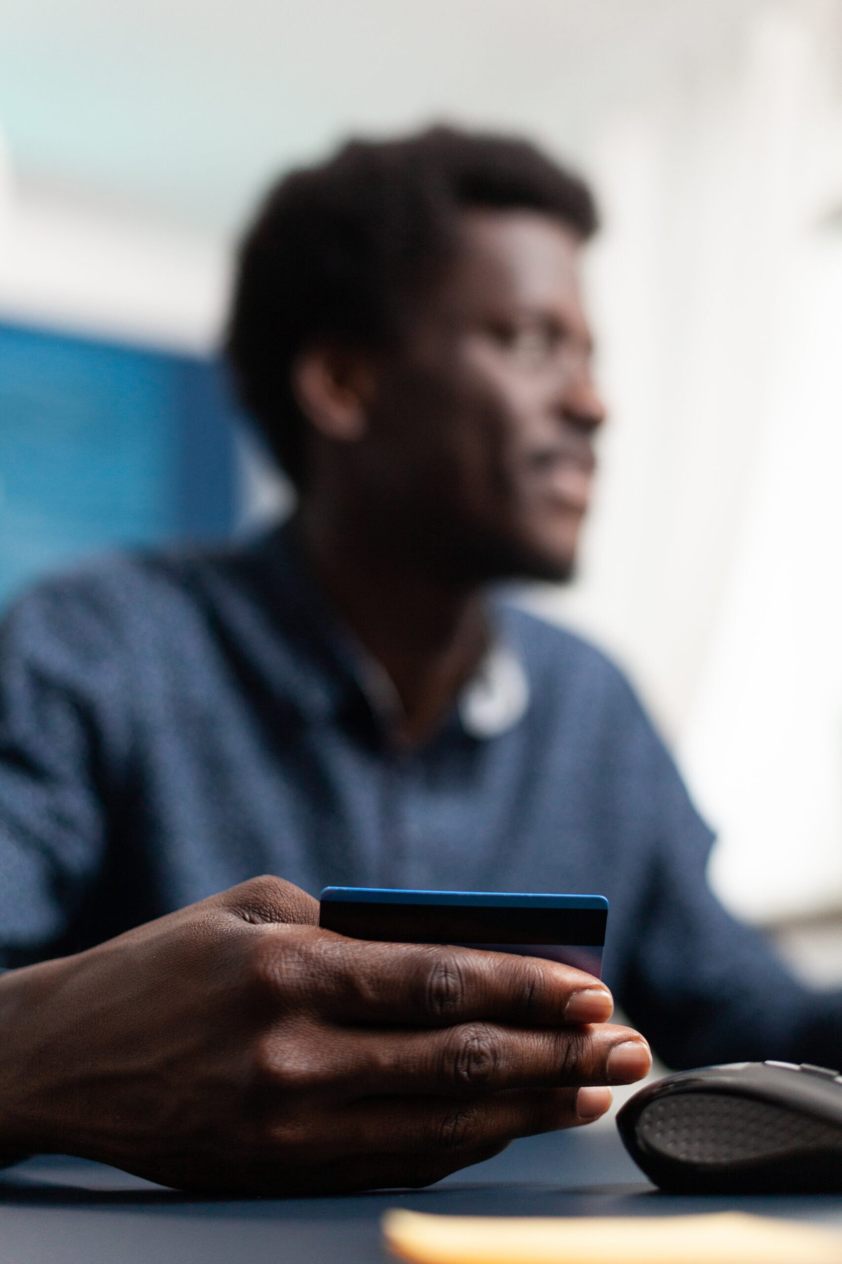 Blurred image of a man with a laptop; the focus is on his hand, holding a credit card. We presume he will make a purchase.